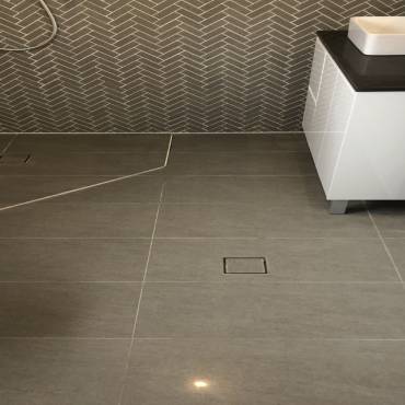 Grey floor tiles cut around a square floor waste beside a vanity